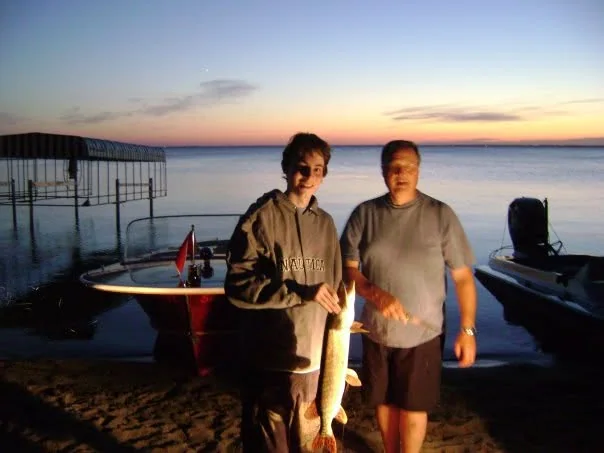 Owen holding a fish beside another person on the shore of Jackfish Lake at sunset